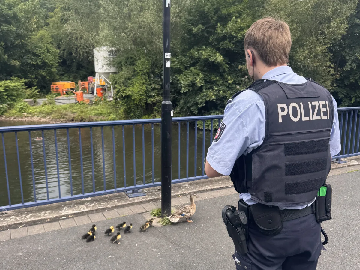 Ein Polizist in Uniform steht auf einer Brücke und beobachtet eine Entenfamilie, die nahe am Fluss entlanggeht.