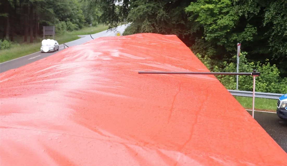 Ein mit einer orangefarbenen Plane bedeckter Lkw steht an einer Landstraße, die von Bäumen gesäumt ist. Neben der Straße ist ein Polizeifahrzeug sichtbar.