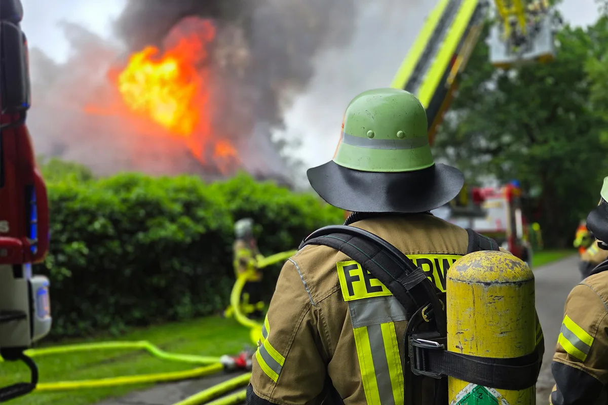 Ein Feuerwehrmann in kompletter Schutzkleidung steht im Vordergrund, während im Hintergrund ein großes Feuer wütet. Mehrere Feuerwehrschläuche sind auf dem Boden verteilt, und Rauch steigt vom Brandherd auf.
