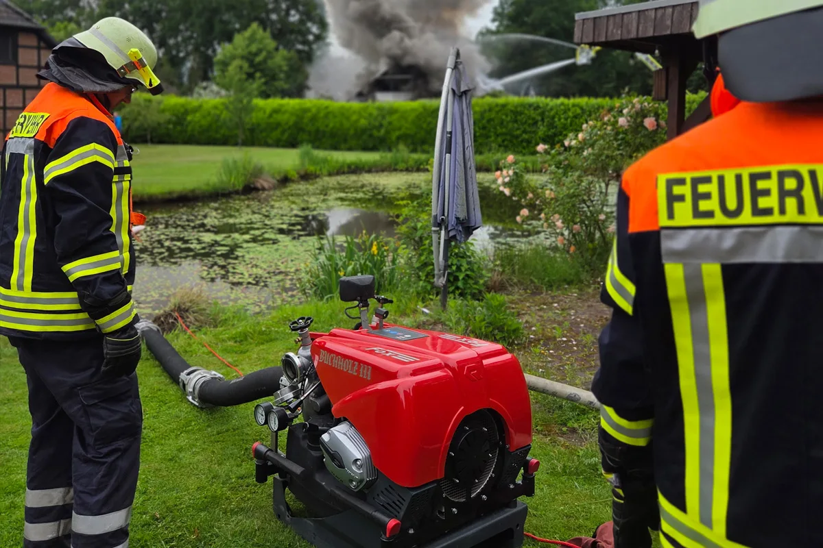 Zwei Feuerwehrleute stehen nahe einer Pumpe vor einem Teich, im Hintergrund ist Rauch von einem Brand zu sehen.