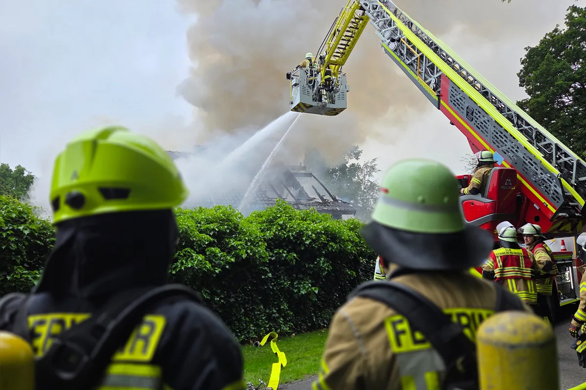Mehrere Feuerwehrleute sind im Einsatz, um mit einer Drehleiter ein brennendes Gebäude zu löschen. Dichter Rauch steigt auf.