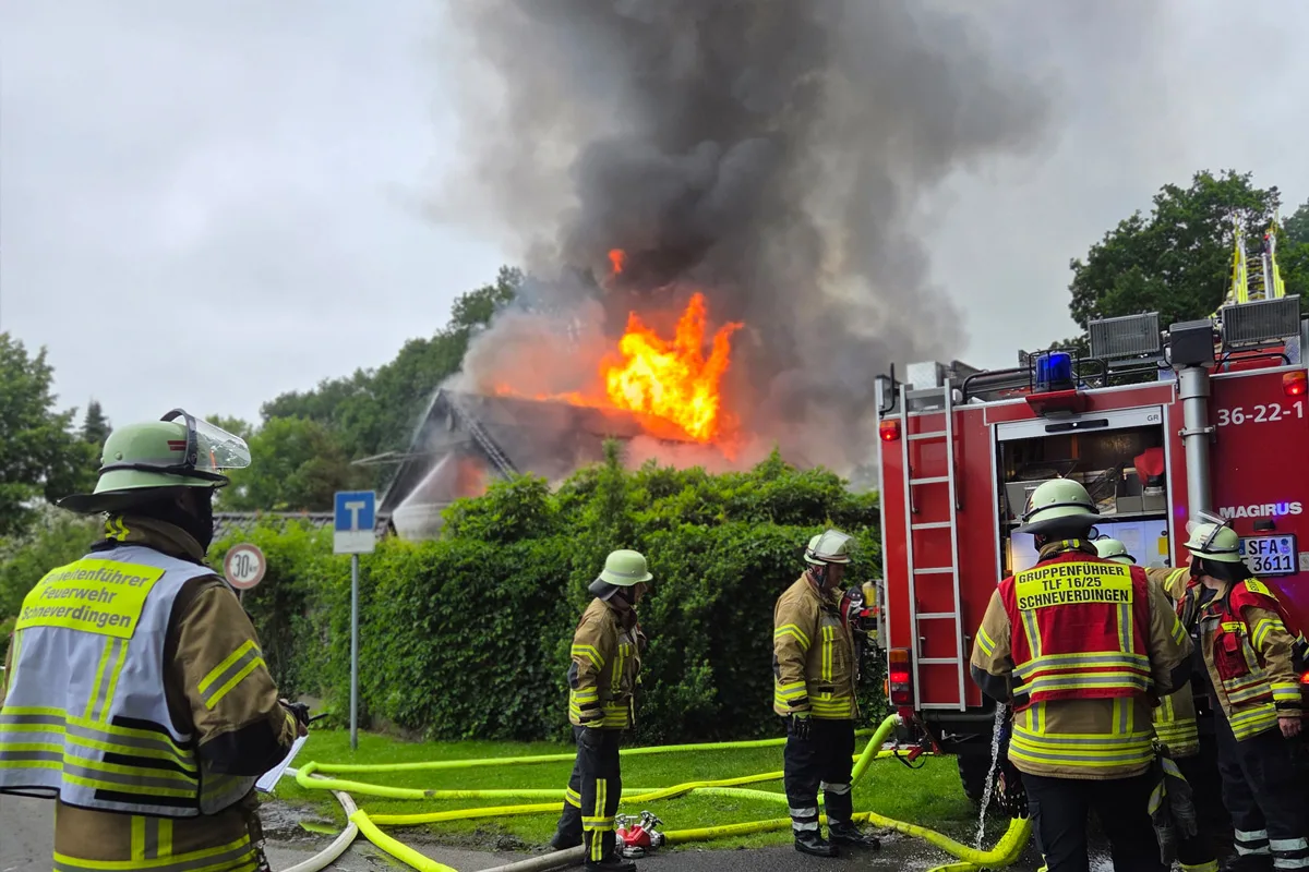Feuerwehrleute stehen vor einem brennenden Haus, aus dessen Dach Flammen schlagen. Ein Feuerwehrfahrzeug ist ebenfalls zu sehen.