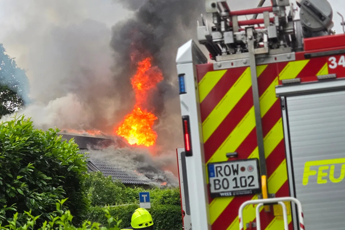 Ein Feuerwehrauto steht vor einem Gebäude, aus dem hohe Flammen und dichter Rauch aufsteigen. Einige Feuerwehrleute sind im Einsatz.
