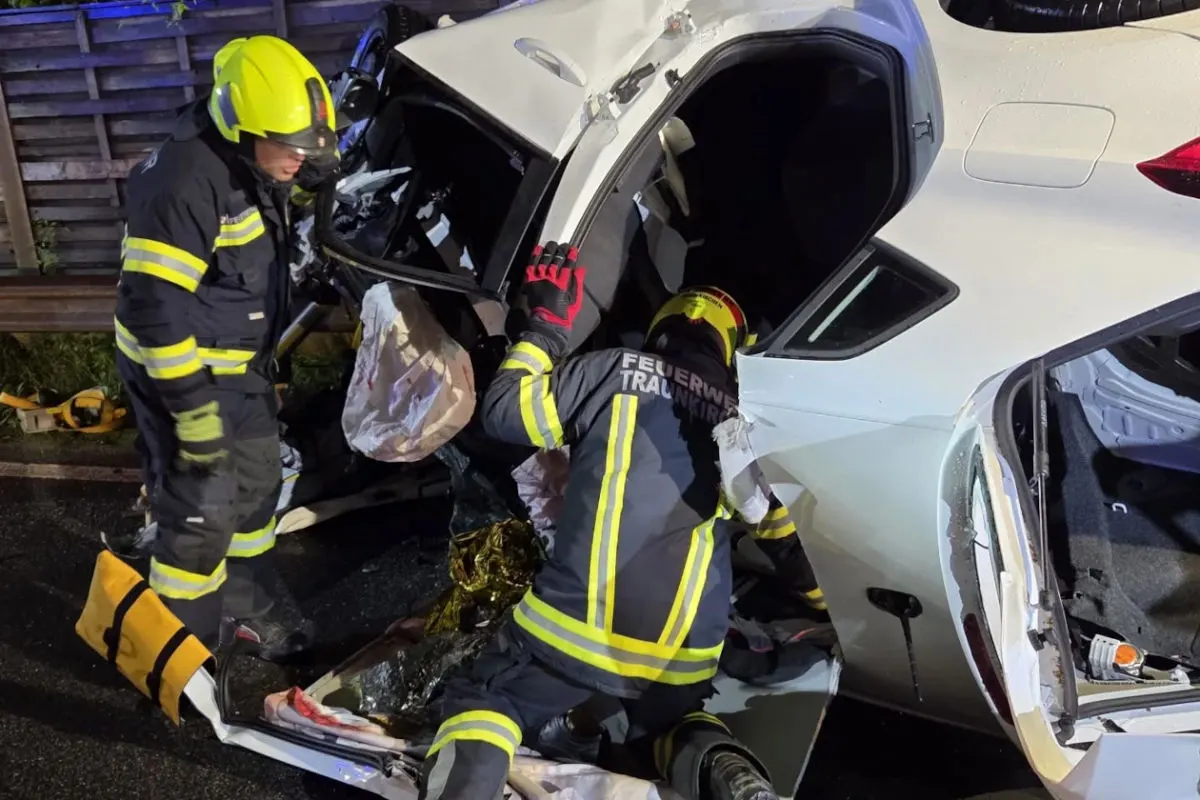 Zwei Feuerwehrleute in Schutzkleidung arbeiten an einem umgestürzten weißen Auto auf der Straße bei Nacht. Das Fahrzeug ist schwer beschädigt.