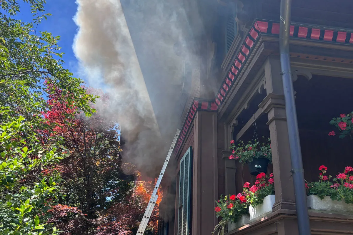 Ein Wohnhaus ist in Brand geraten, Rauch und Flammen steigen auf. Blumen sind auf dem Balkon sichtbar.