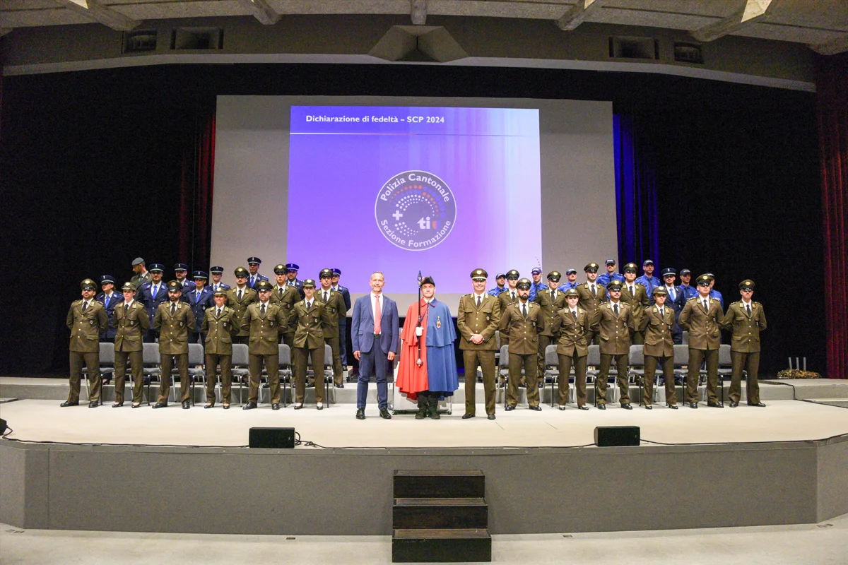 Mehrere uniformierte Personen posieren auf einer Bühne vor einer Leinwand mit einer Projektion. Die Uniformen sind vorwiegend grün.