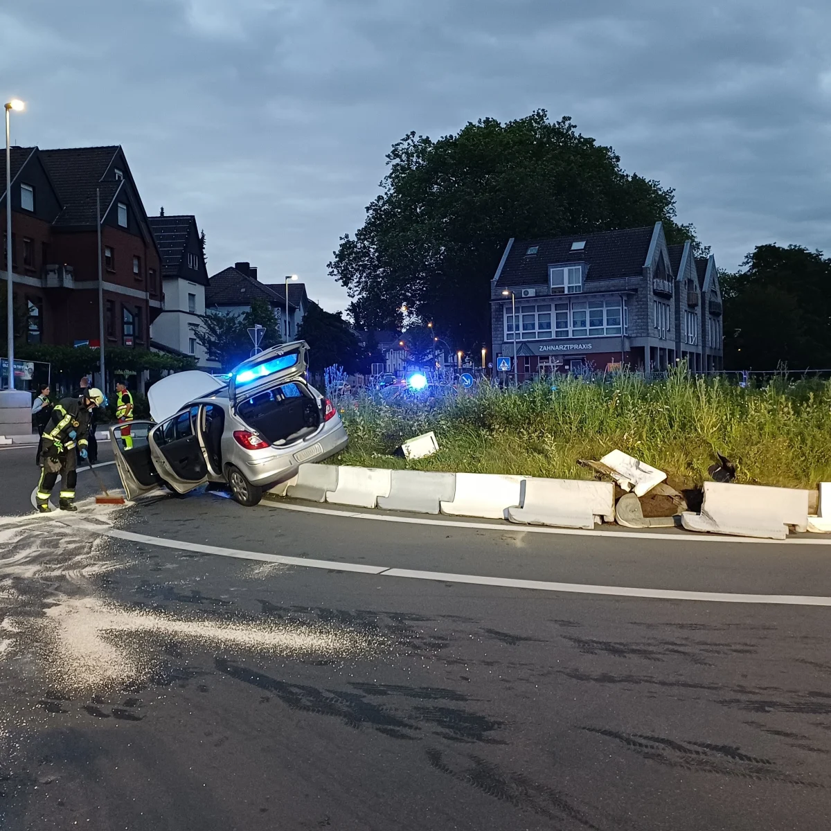 Ein Auto hängt schräg auf einer Straßenbarriere neben einer Verkehrsinsel. Einsatzkräfte sind vor Ort, zu sehen bei Nachtlicht in der Stadt.