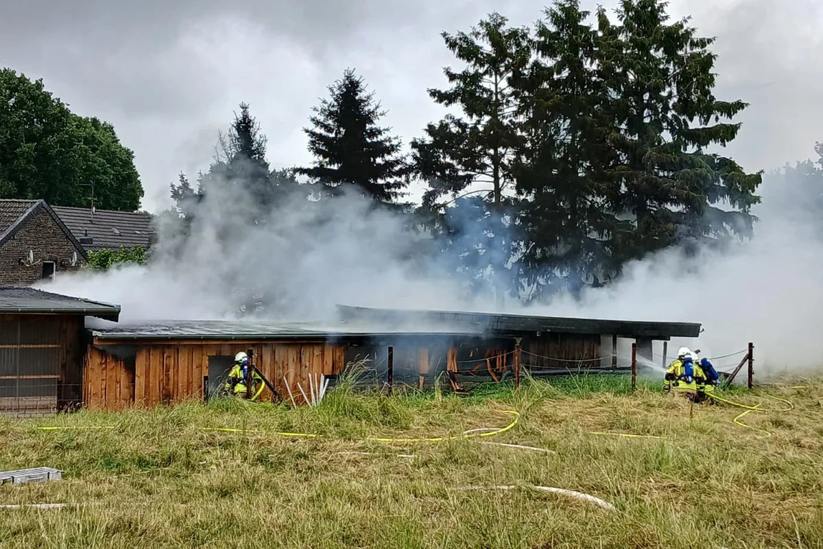 Zwei Feuerwehrleute bekämpfen einen Brand in einem Schuppen auf einem Feld, während Rauch aufsteigt. Im Hintergrund sind Bäume und Häuser zu sehen.