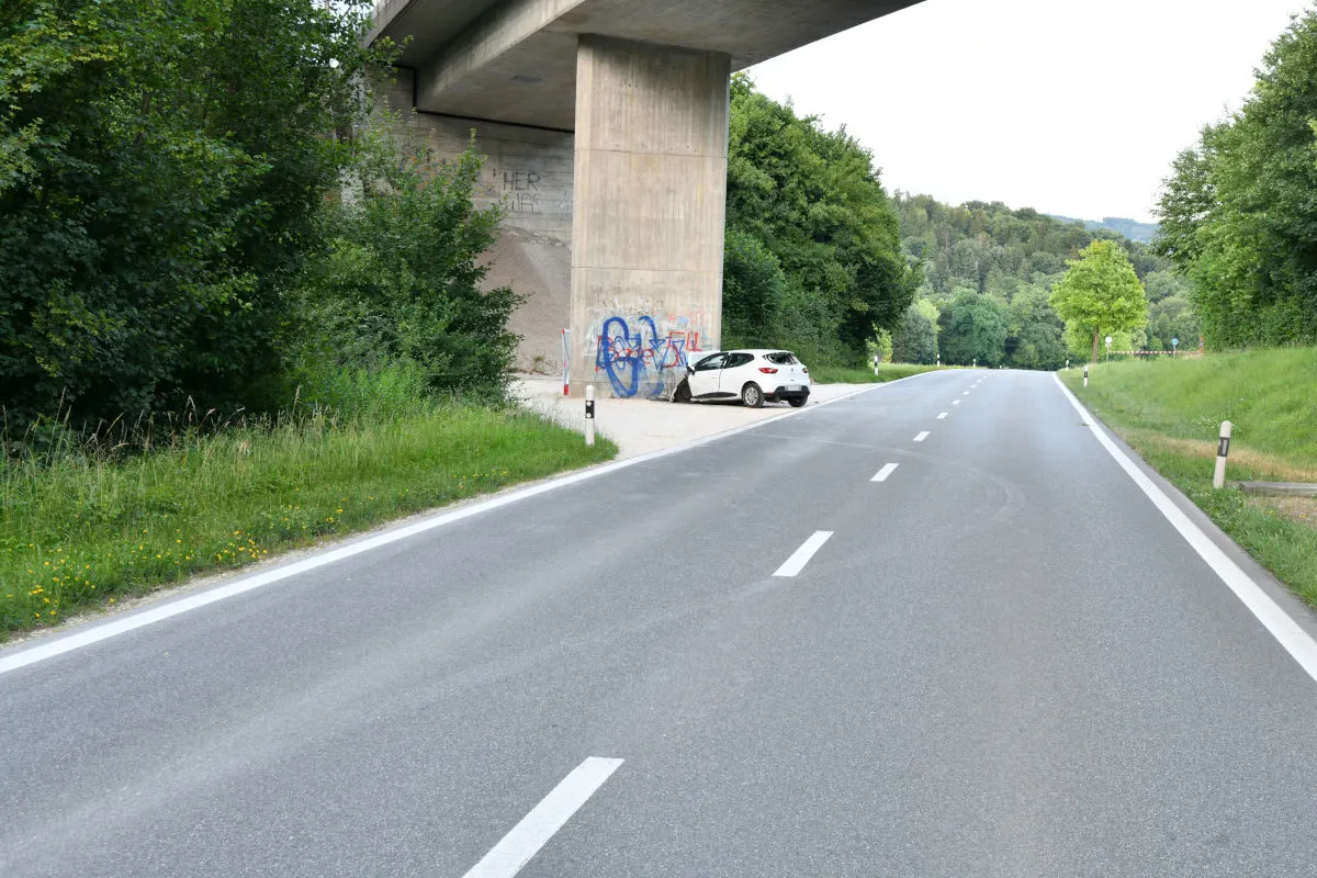 Ein weißes Auto ist unter eine Brücke gefahren und zeigt eine sichtbare Seitenbeschädigung. Der Standort ist von grüner Vegetation umgeben.