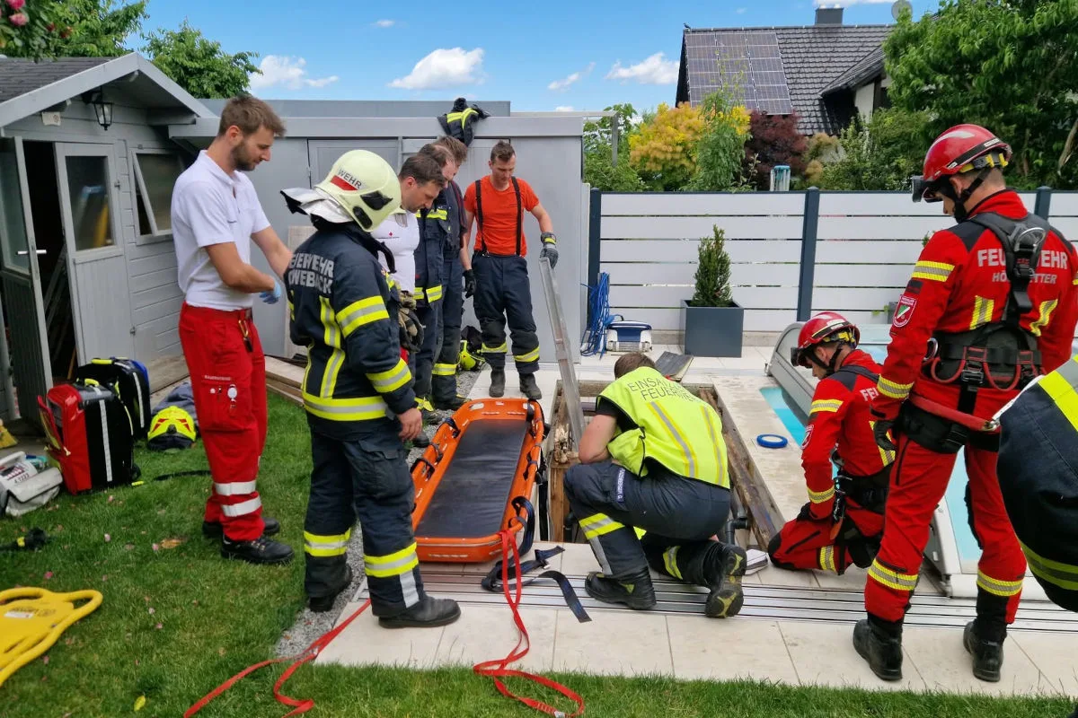 Mehrere Rettungskräfte in unterschiedlichen Uniformen arbeiten an einer offenen Poolabdeckung neben einem Rasenstück. Ein Notarbeitskoffer ist in der Nähe.