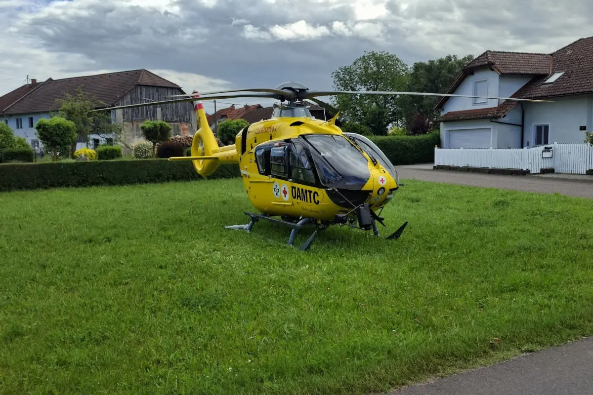 Ein gelber Rettungshubschrauber des ÖAMTC ist auf einer grünen Wiese in einem Wohngebiet gelandet. Im Hintergrund sind Wohnhäuser zu sehen.