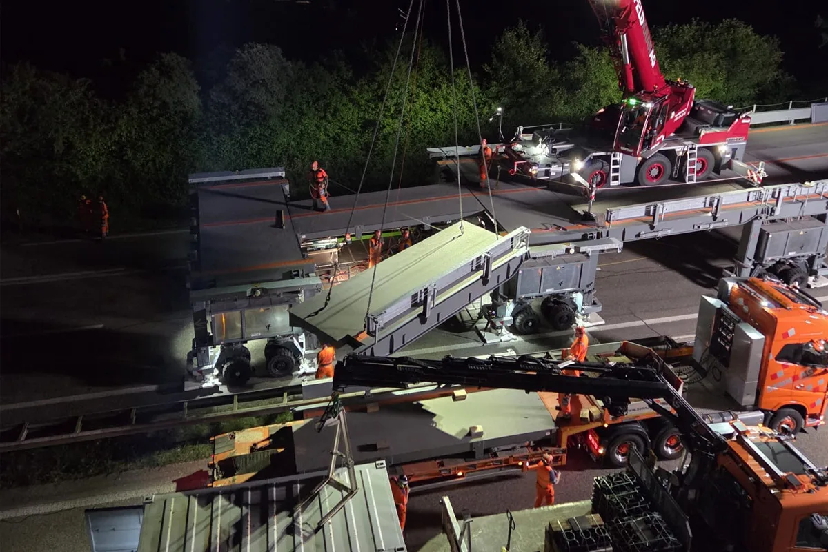 Ein Kran bewegt bei Nacht ein großes Brückenteil, Arbeiter stehen neben den Fahrzeugen auf der Autobahn.