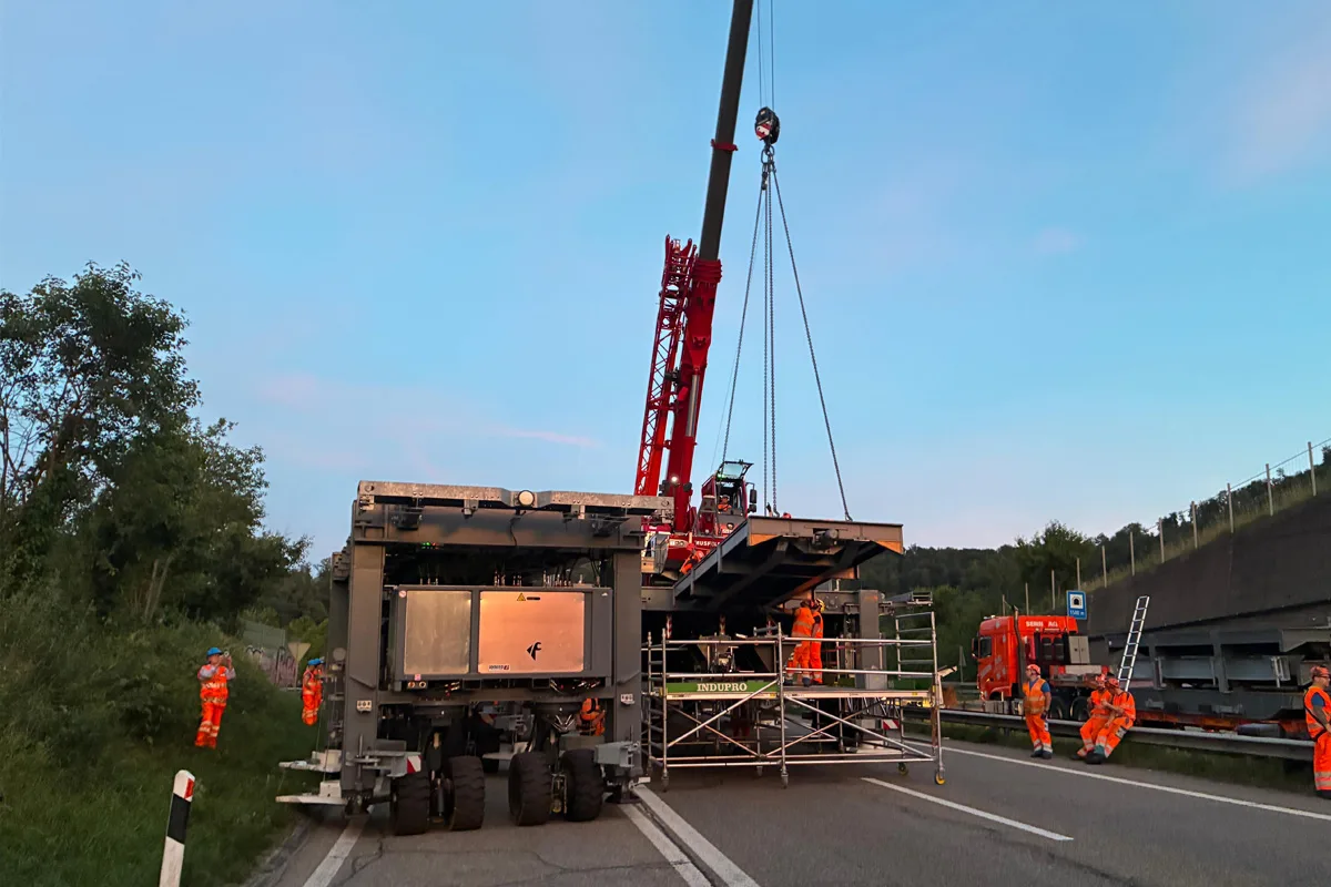 Ein großer Kran hebt eine Metallkonstruktion von einem Schwerlasttransporter. Mehrere Arbeiter in orangefarbener Schutzkleidung sind tätig, während die Szenerie im Abendlicht stattfindet.