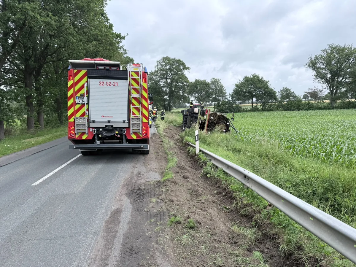 Ein umgekippter Holztransporter liegt neben einer Landstraße an einem Feld. Im Hintergrund sind Feuerwehrleute sichtbar, die die Situation sichern.
