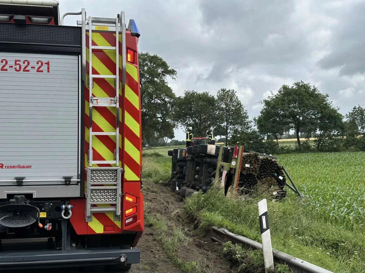 Ein Feuerwehrfahrzeug steht am Rand einer Straße, während ein umgekippter Lkw voll mit Holzstämmen auf einer Wiese liegt. Zwei Feuerwehrleute sind auf dem Lkw zu sehen.