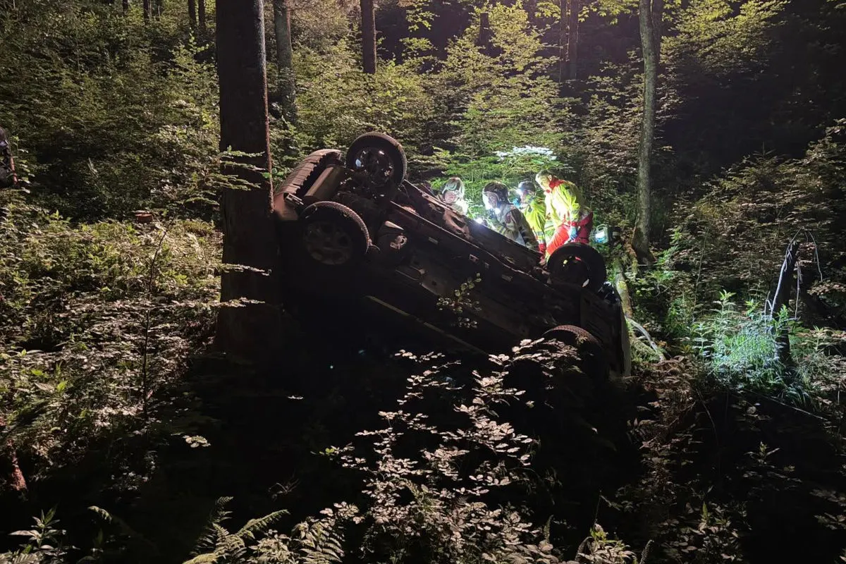 Ein Auto liegt bei Nacht kopfüber in einem dichten Wald. Mehrere Rettungskräfte stehen mit Scheinwerfern um das Fahrzeug herum.