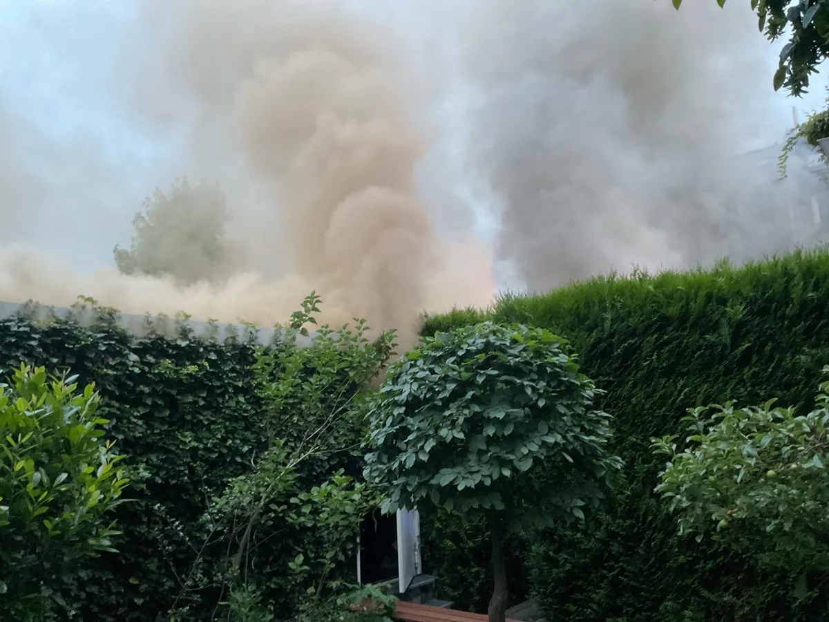 Dichte Rauchwolken steigen hinter hohen, grünen Hecken auf; es ist keine Person oder Fahrzeug sichtbar.