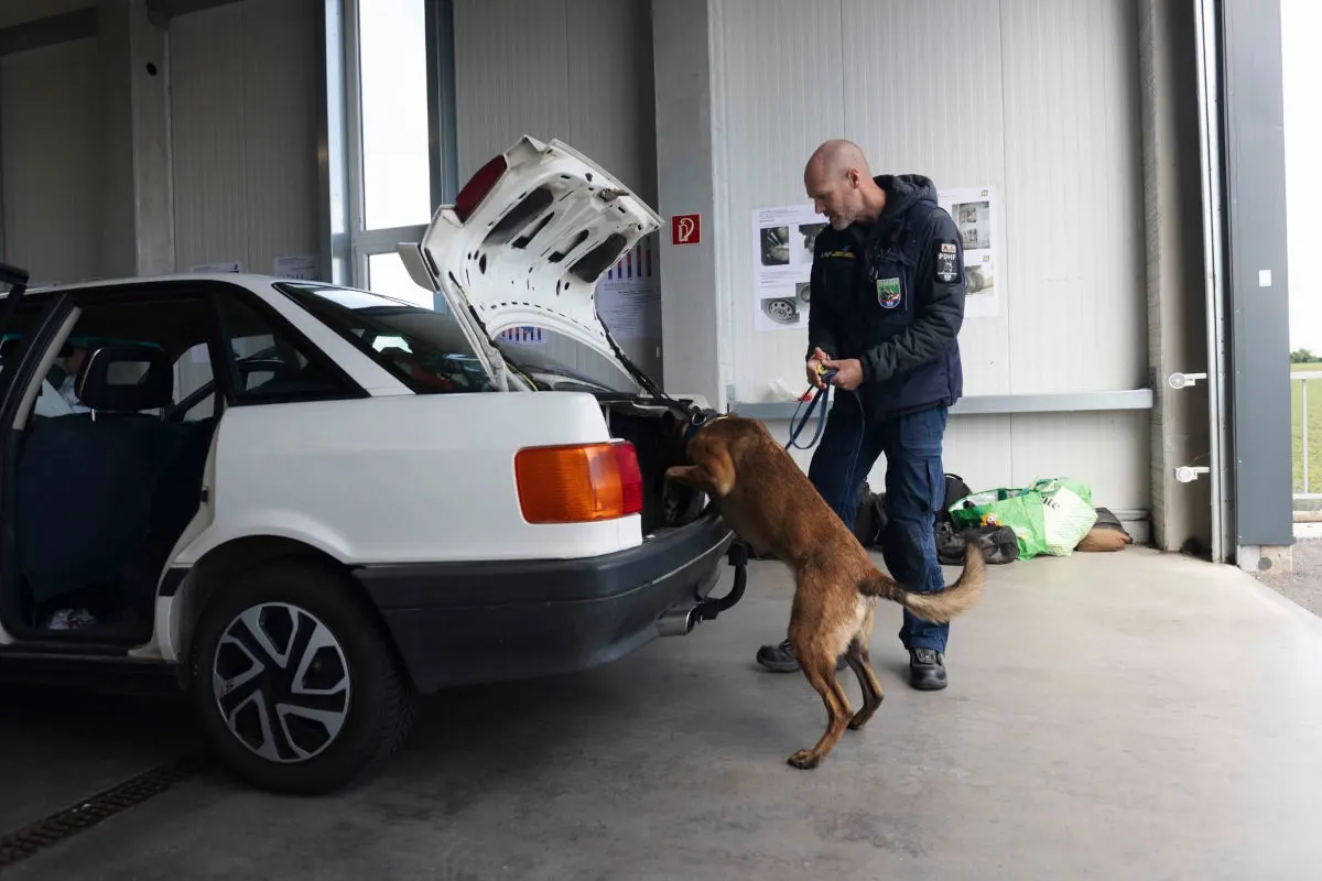 Ein Polizist mit einem Spürhund untersucht den Kofferraum eines weißen Autos in einer Halle. Der Hund schnüffelt aktiv im offenen Kofferraum, während der Beamte aufmerksam zusieht.