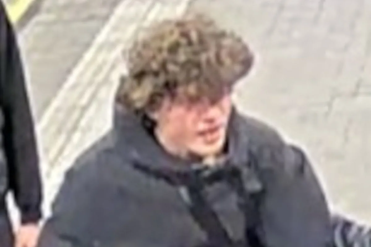 Ein junger Mann mit lockigem Haar trägt dunkle Kleidung und steht auf einem Gehweg. Der Hintergrund ist unscharf.
