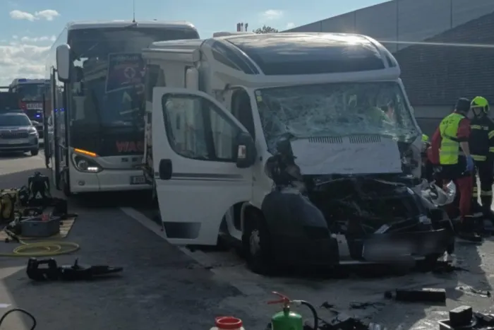 Das Bild zeigt ein beschädigtes Wohnmobil mit zerbrochener Windschutzscheibe und geöffneter Tür auf einer Autobahn. Im Hintergrund sind ein Bus und Einsatzkräfte sichtbar.