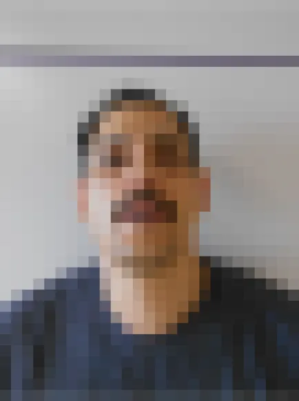 Ein Mann mit kurzen Haaren und Schnurrbart blickt in die Kamera vor einem neutralen Hintergrund.