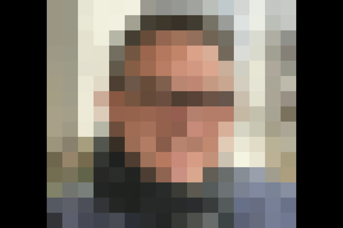 Ein Mann mit Brille und Schal lächelt in die Kamera. Der Hintergrund ist unscharf und zeigt Pflanzen und eine Mauer.