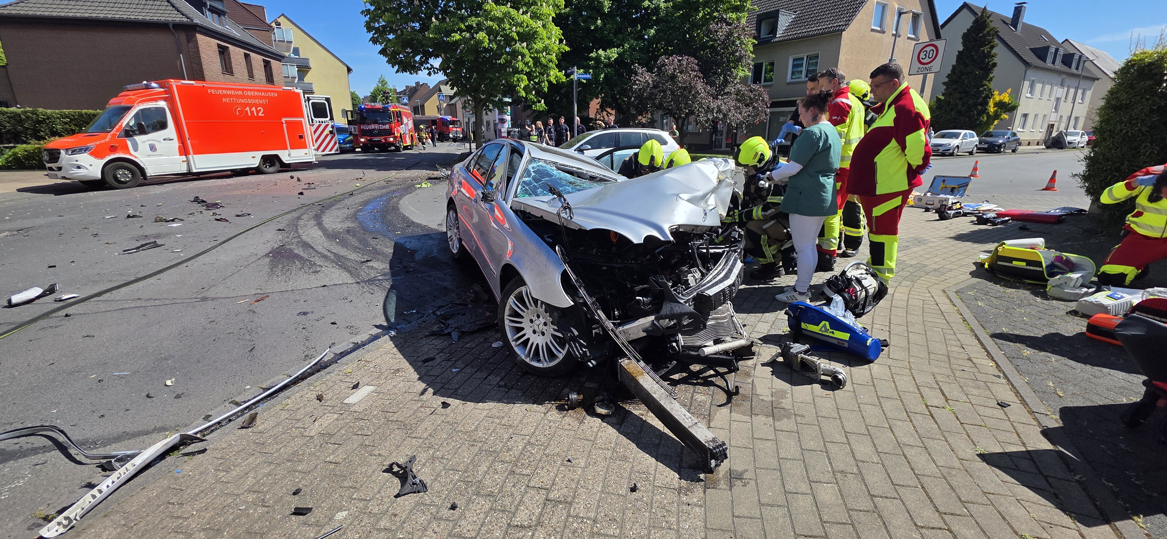 Ein silbernes Auto mit stark beschädigter Vorderseite steht auf einer Straße. Ringsherum sind Feuerwehr- und Rettungskräfte im Einsatz. Im Hintergrund ist ein Feuerwehrfahrzeug zu sehen.