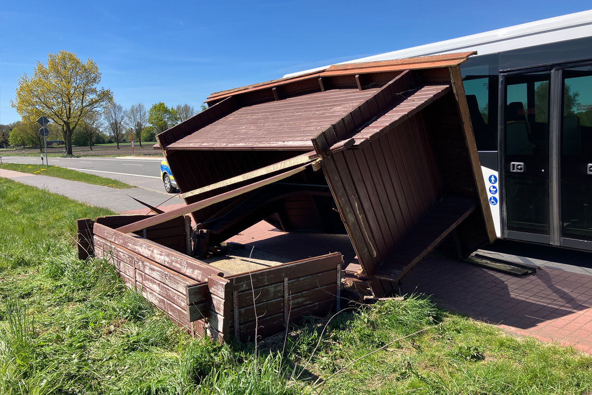 Eine beschädigte Holzhütte liegt umgekippt an einem Bus auf einer Landstraße. Der Himmel ist klar und die Bäume sind grün.