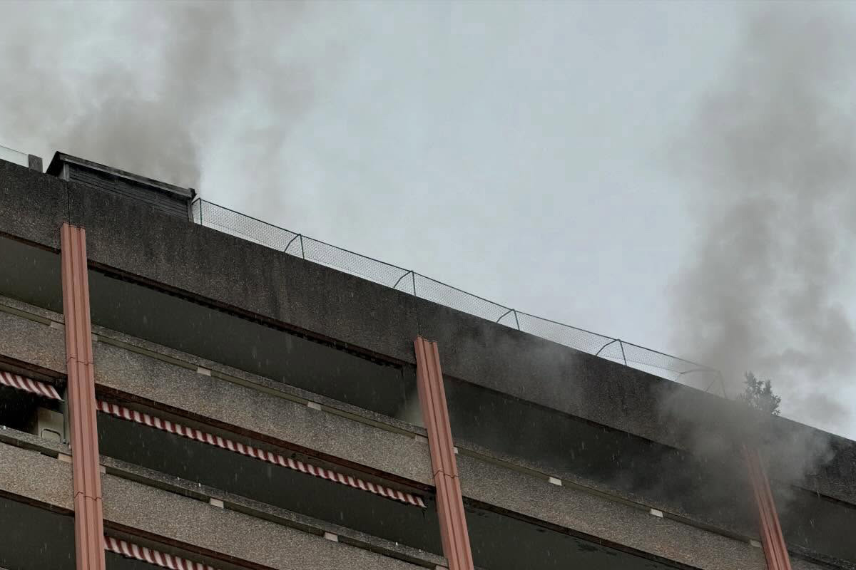 Ein mehrstöckiges Gebäude mit Flachdach, von dem an mehreren Stellen Rauch aufsteigt. Es ist bewölkt.