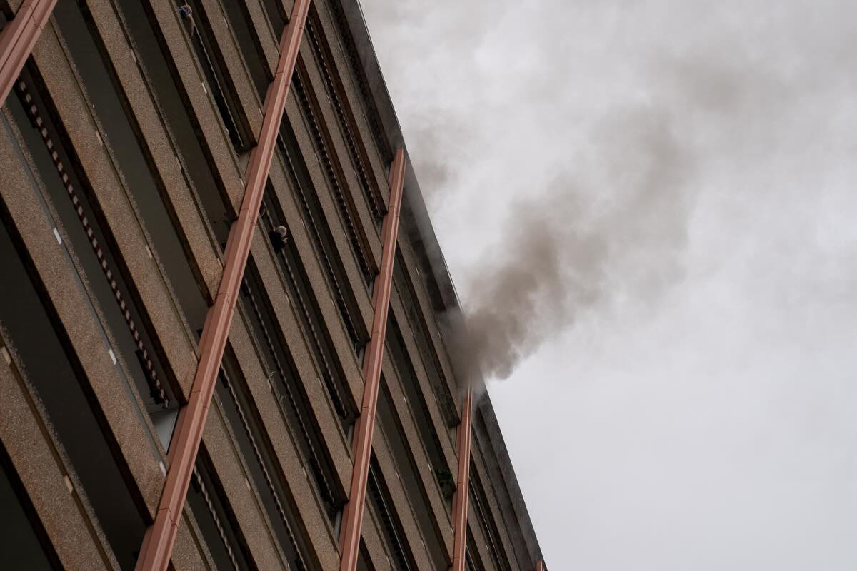 Ein mehrstöckiges Gebäude mit dichtem Rauch, der aus einem Fenster in den grauen Himmel strömt, ist sichtbar.