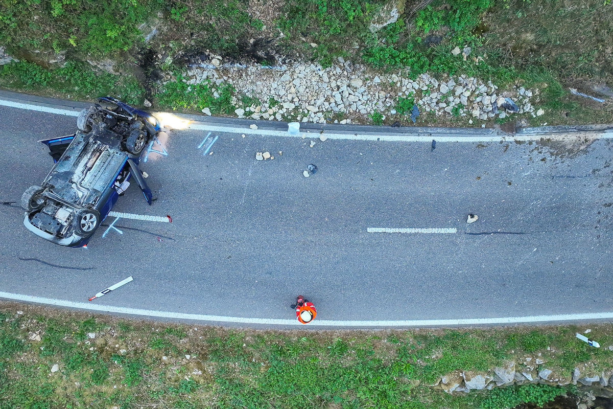 Ein Auto liegt auf dem Dach auf einer zweispurigen Straße, umgeben von Trümmern. Eine Person in Warnweste steht daneben.