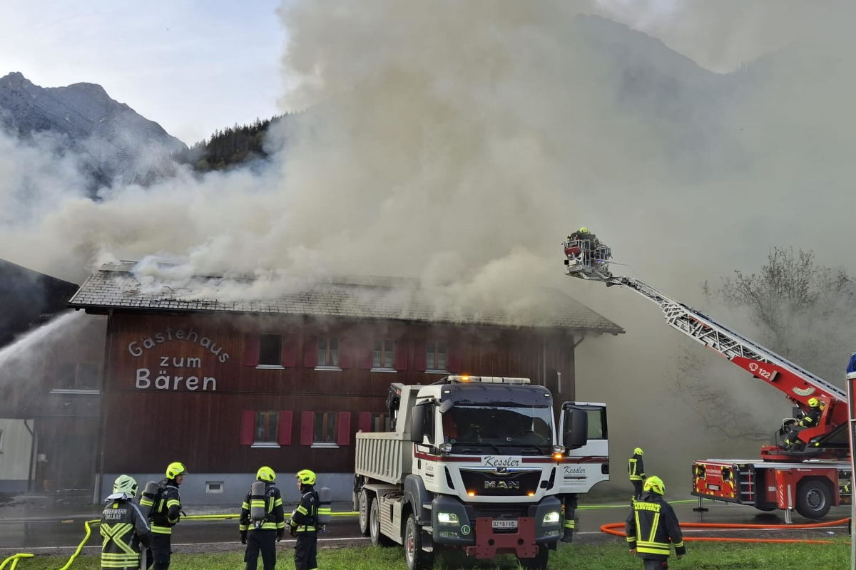Mehrere Feuerwehrleute sind zu sehen, die einen Brand in einem Holzgebäude bekämpfen. Dichte Rauchwolken steigen auf und ein Feuerwehrfahrzeug ist im Einsatz.
