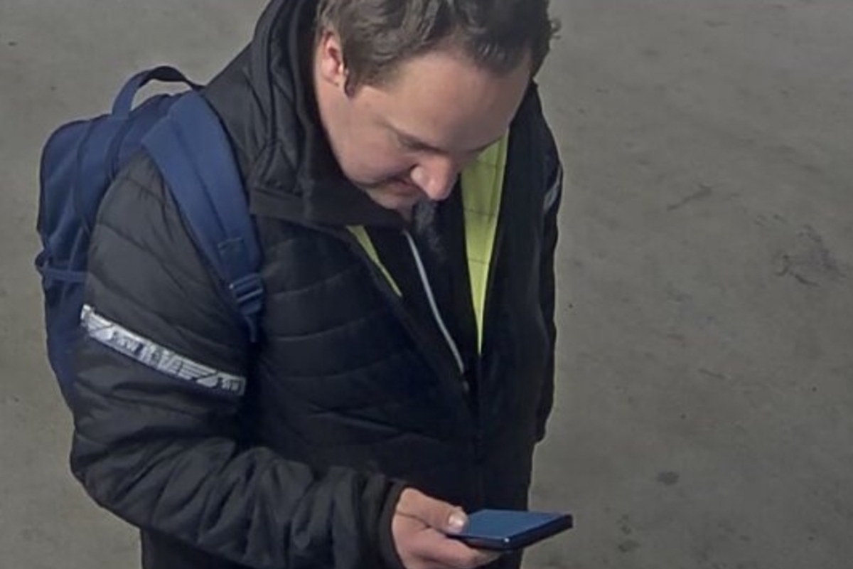 Eine Person steht in einem Innenbereich, trägt eine dunkle Jacke und einen blauen Rucksack, und blickt auf ein Smartphone in der Hand.