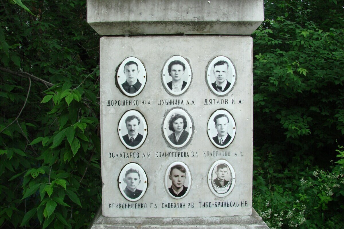 Eine Steinsäule mit neun ovalen Portraits, daneben dichter Wald. Die Portraits sind in drei Reihen angeordnet.