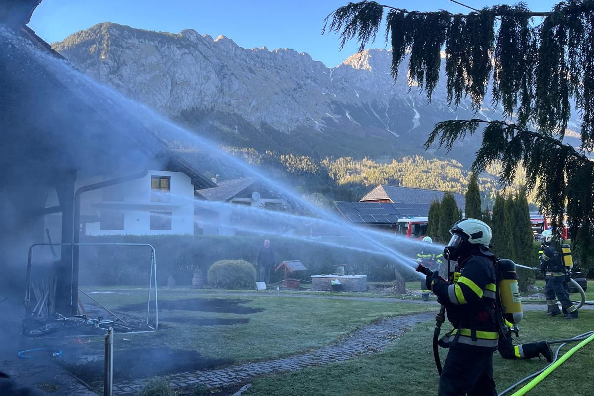 Im Vordergrund sind Feuerwehrleute mit Atemschutzgeräten zu sehen, die Wasser auf ein Gebäude spritzen. Im Hintergrund erhebt sich eine Berglandschaft unter einem blauen Himmel.