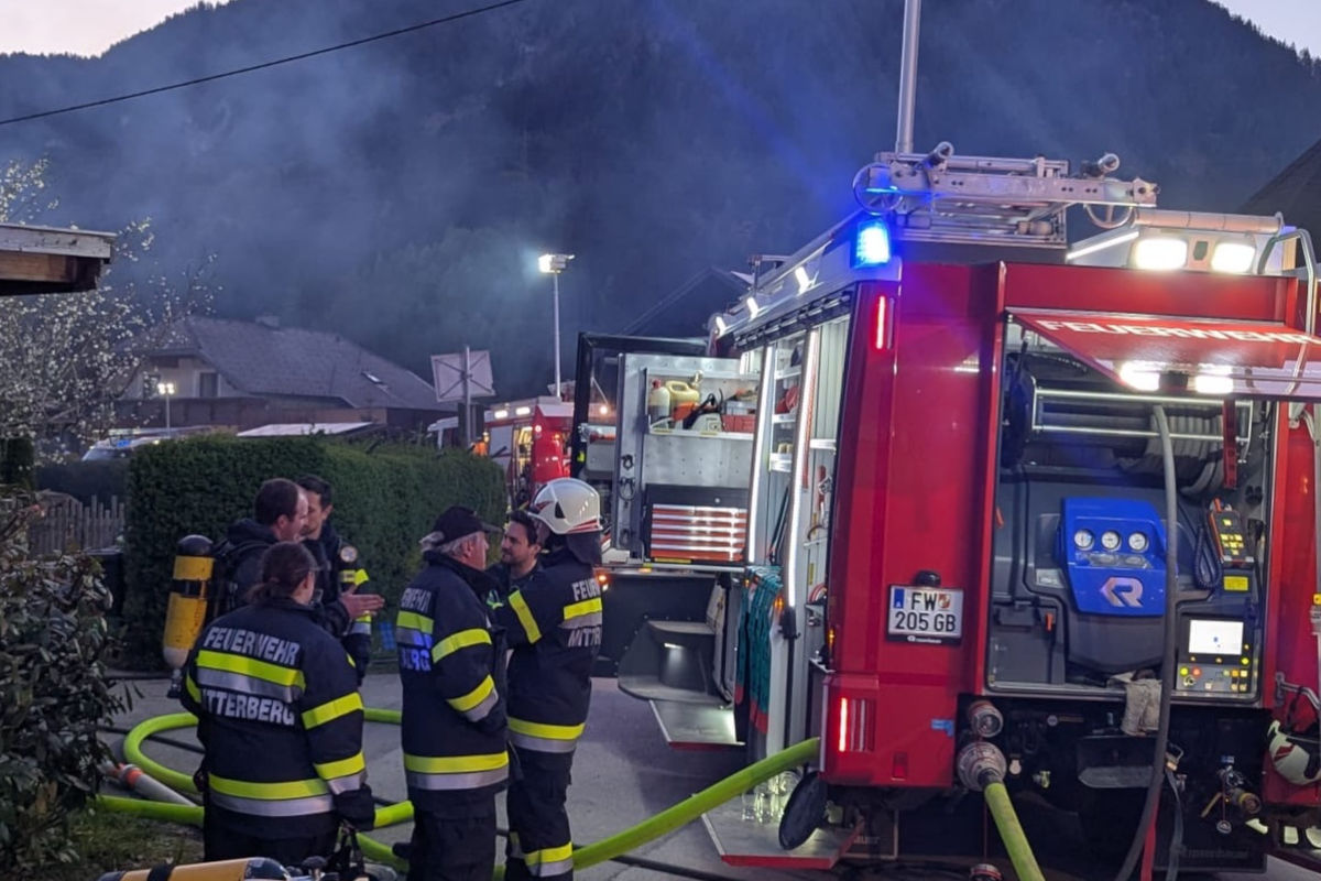 Mehrere Feuerwehrleute stehen in einer ruhigen Wohngegend bei einem roten Feuerwehrauto und unterhalten sich. Im Hintergrund sind Berge und dämmerndes Licht sichtbar.