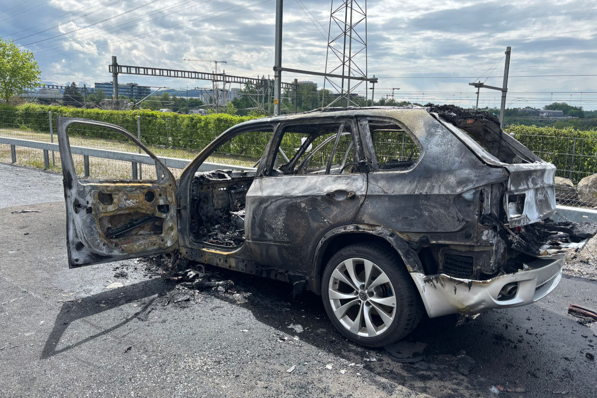 Ein vollständig ausgebranntes Auto steht mit geöffneter Tür auf einer Straße. Im Hintergrund sind Bahngleise und Infrastruktur zu sehen.
