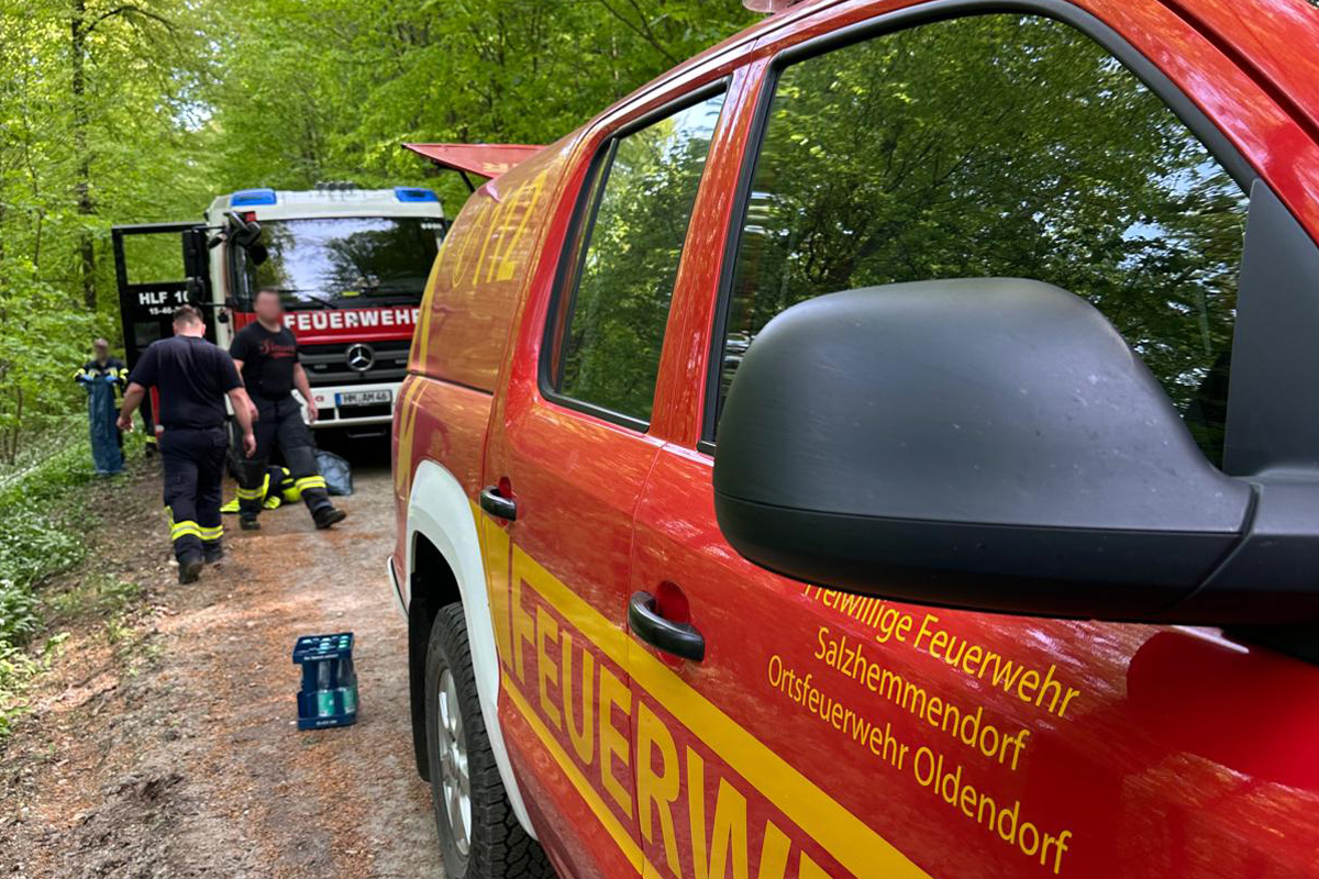 Zwei Feuerwehrfahrzeuge stehen auf einem Waldweg, umgeben von grünen Bäumen. Mehrere Feuerwehrleute in Uniform sind zu sehen.