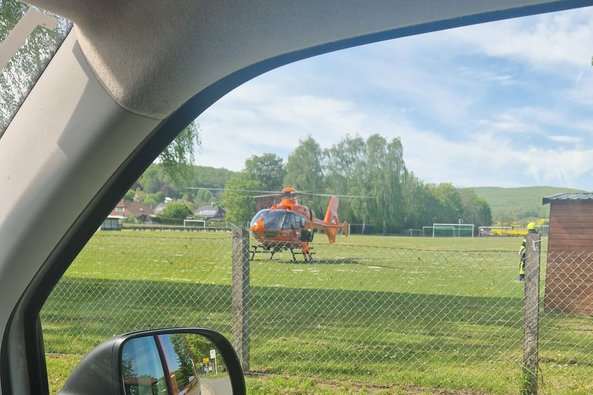 Durch das Autofenster sieht man einen orangefarbenen Hubschrauber auf einem umzäunten Rasenplatz stehen. Ein Feuerwehrmann ist in der Nähe des Hubschraubers sichtbar.
