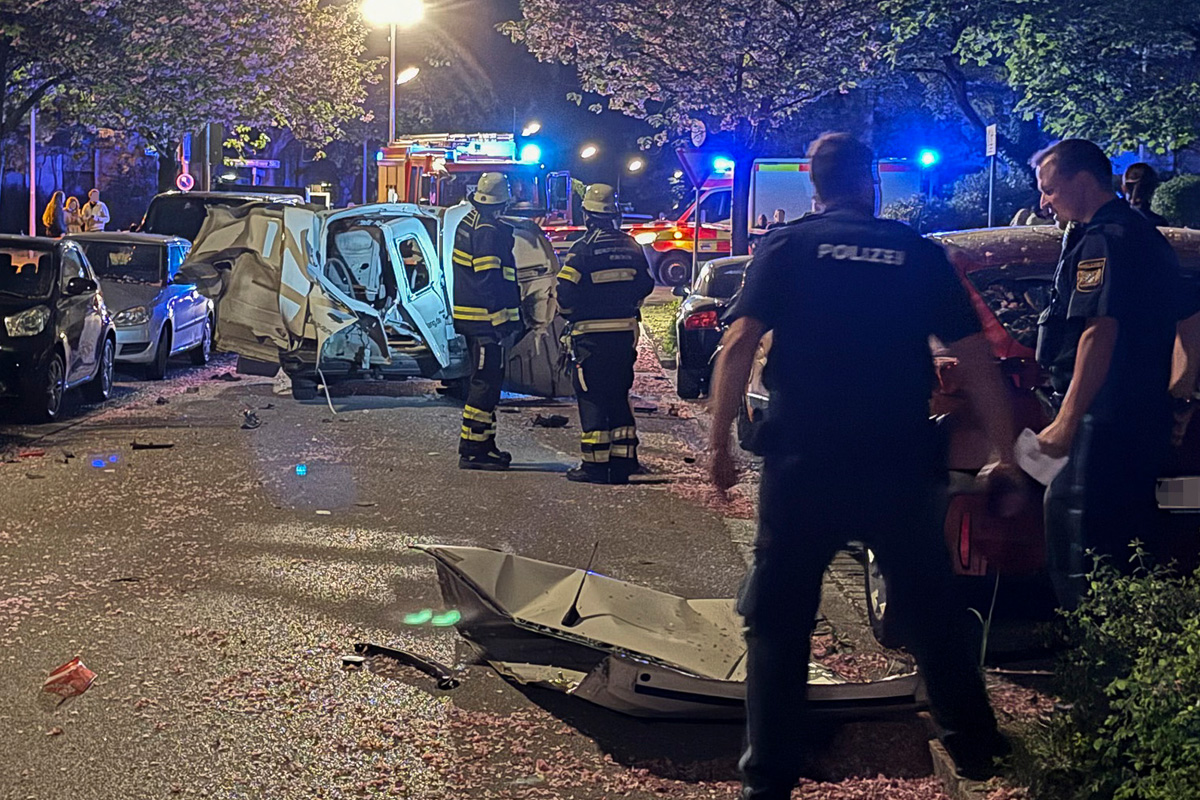 Auf einer nächtlichen Straße befindet sich ein schwer beschädigtes Fahrzeug. Einige Einsatzkräfte in Uniform stehen in der Nähe, während Polizei und Feuerwehr vor Ort sind.