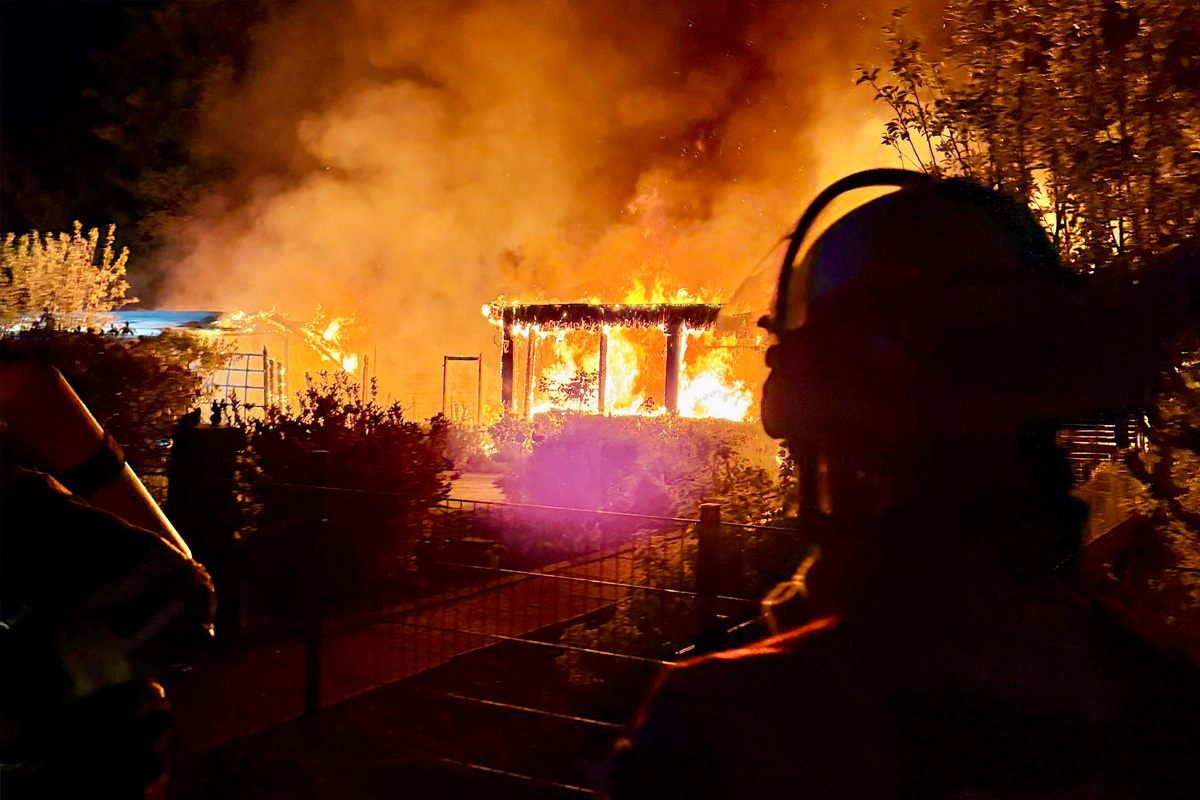 Ein Gebäude steht in Flammen bei Nacht. Im Vordergrund sind die Silhouetten von Feuerwehrleuten zu erkennen, die die Szene beobachten.