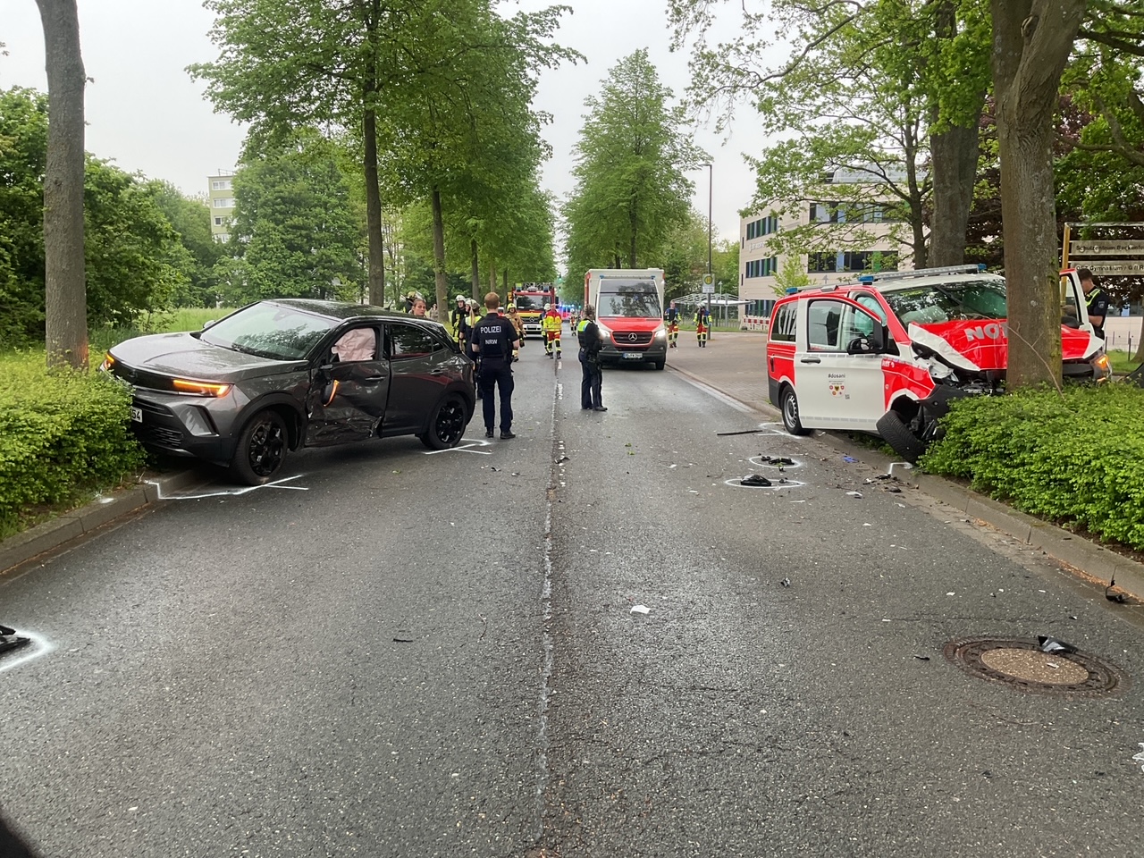 Ein graues Auto und ein rot-weißes Notarztfahrzeug sind in einen Unfall verwickelt. Auf der Straße stehen mehrere Personen, darunter Polizisten.
