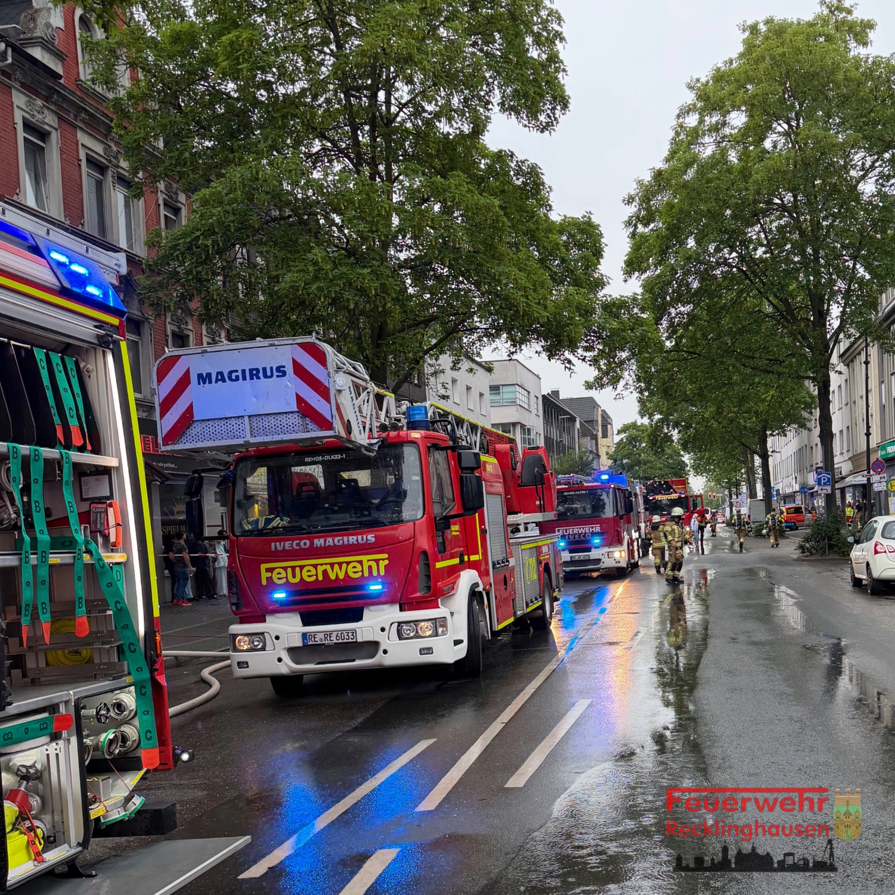 Mehrere rote Feuerwehrfahrzeuge stehen auf einer regennassen Straße mit Bäumen und Gebäuden im Hintergrund. Feuerwehrleute sind bei den Fahrzeugen zu sehen.