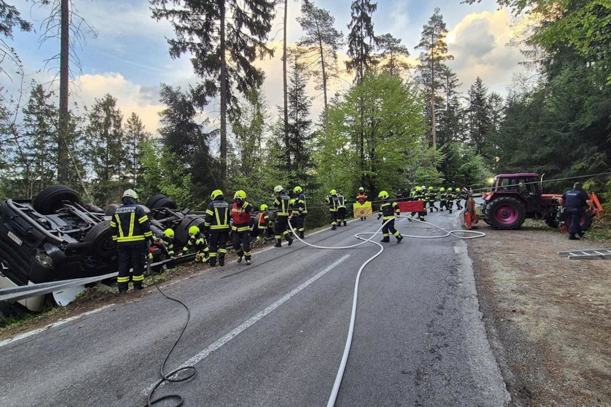 Eine Gruppe von Feuerwehrleuten in leuchtender Schutzkleidung arbeitet an einem umgestürzten Fahrzeug, das auf einer Waldstraße liegt. Im Hintergrund sind Bäume und ein Himmel mit ein paar Wolken zu sehen.