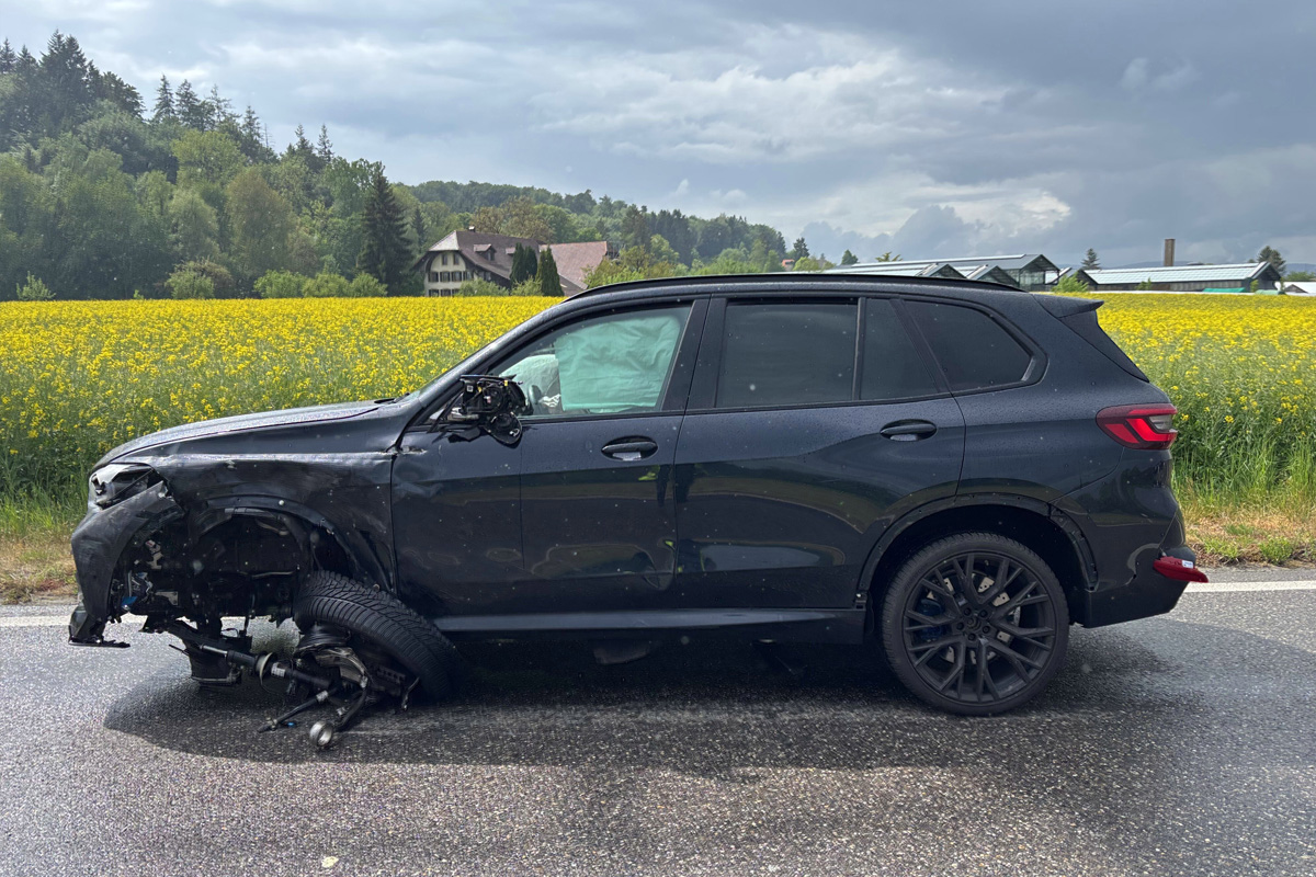 Ein schwarzes Auto mit schweren Frontschäden steht am Straßenrand. Im Hintergrund sind Felder und Häuser zu sehen.