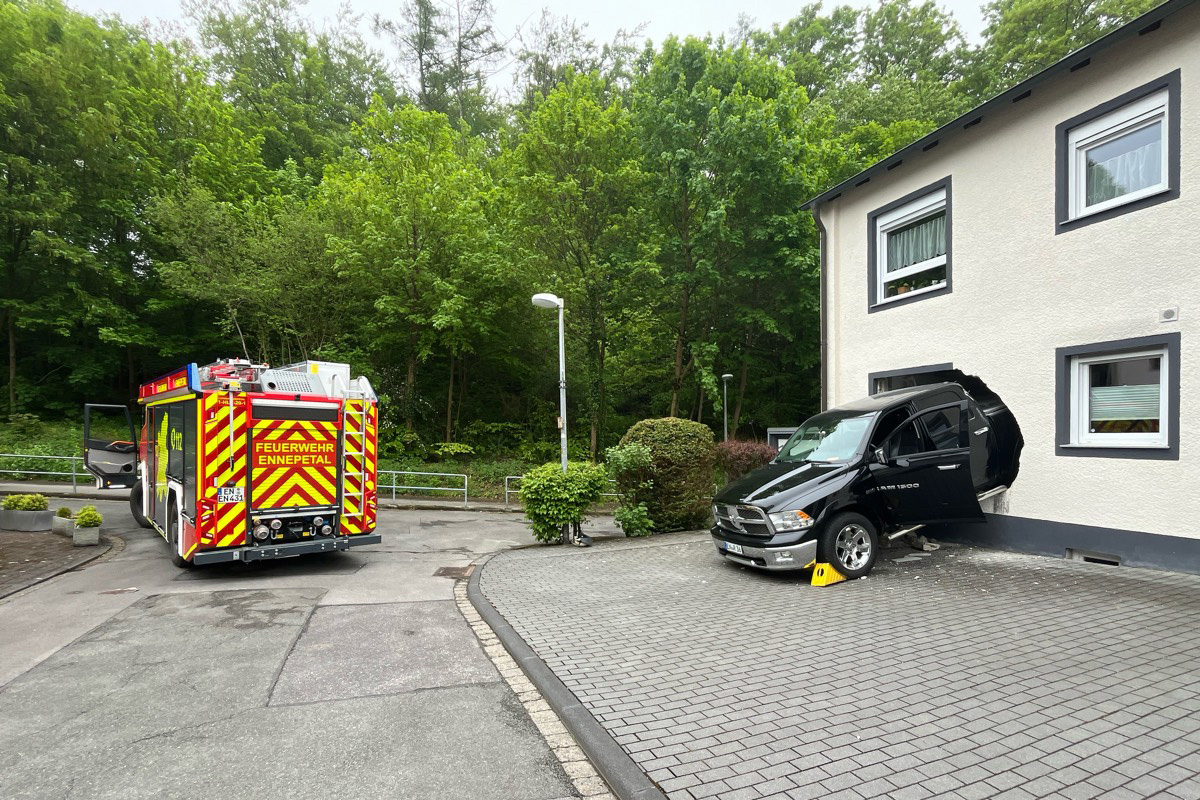 Ein schwarzes Auto ist teilweise in eine beige Hauswand gefahren. Daneben steht ein Feuerwehrfahrzeug mit der Aufschrift 'Feuerwehr Ennepetal'.