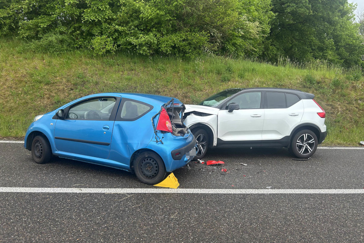 Ein blauer Kleinwagen mit beschädigtem Heck und ein weißer SUV mit beschädigter Front stehen auf einer Straße am Straßenrand. Der Hintergrund ist grün mit Bäumen und Gras.