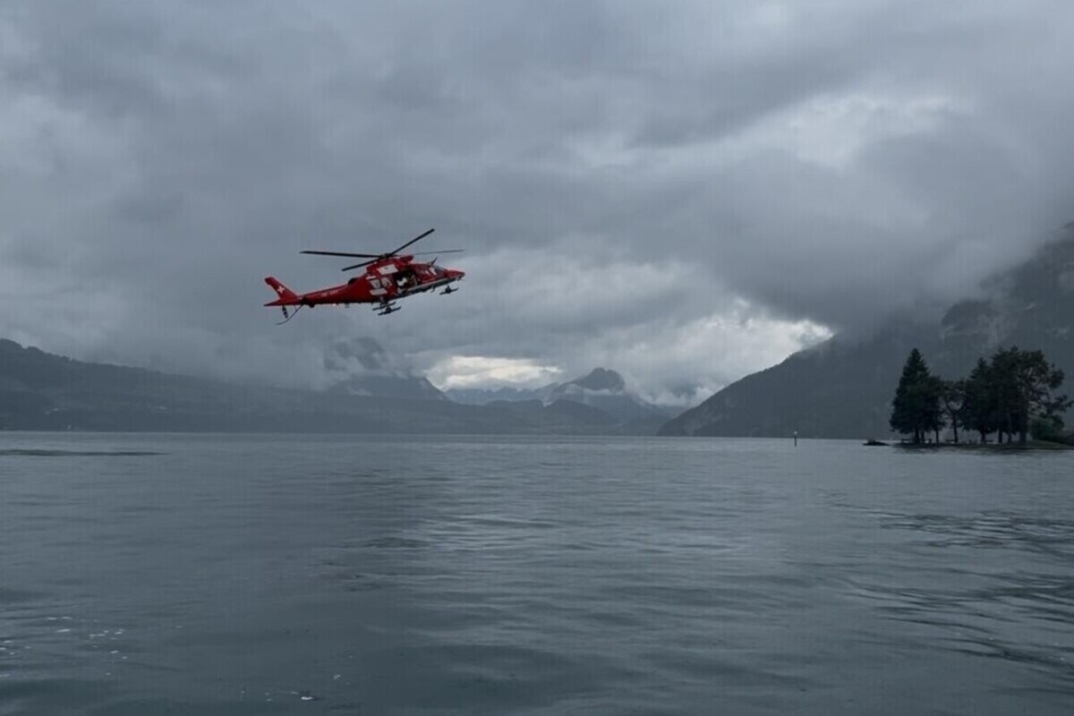 Ein roter Rettungshubschrauber schwebt über einem ruhigen See unter einem bewölkten Himmel. Im Hintergrund sind Berge sichtbar.