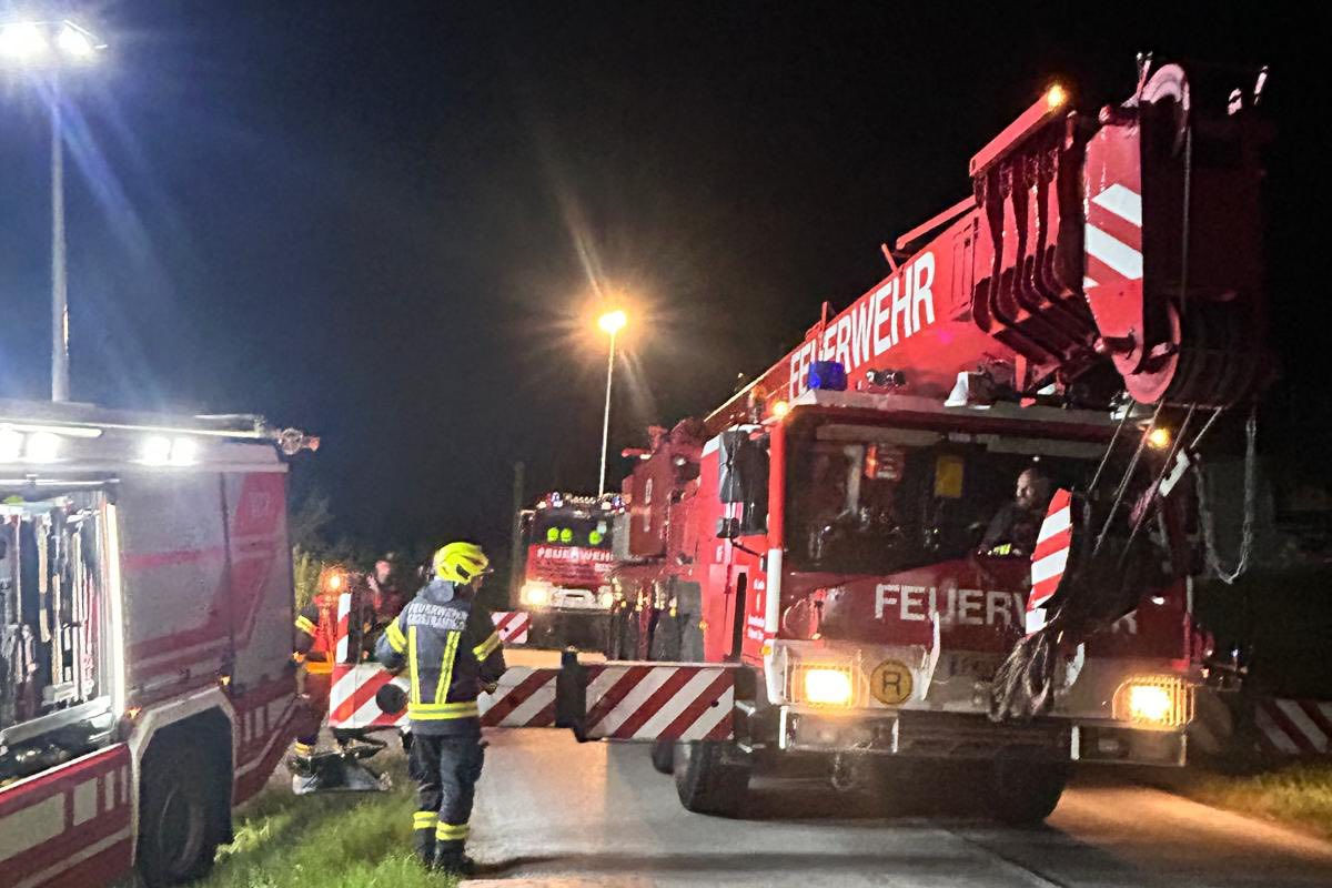 Mehrere Feuerwehrfahrzeuge sind auf einer nächtlichen Straße im Einsatz. Einsatzkräfte in gelben Schutzhelmen sind ebenfalls vor Ort zu sehen.