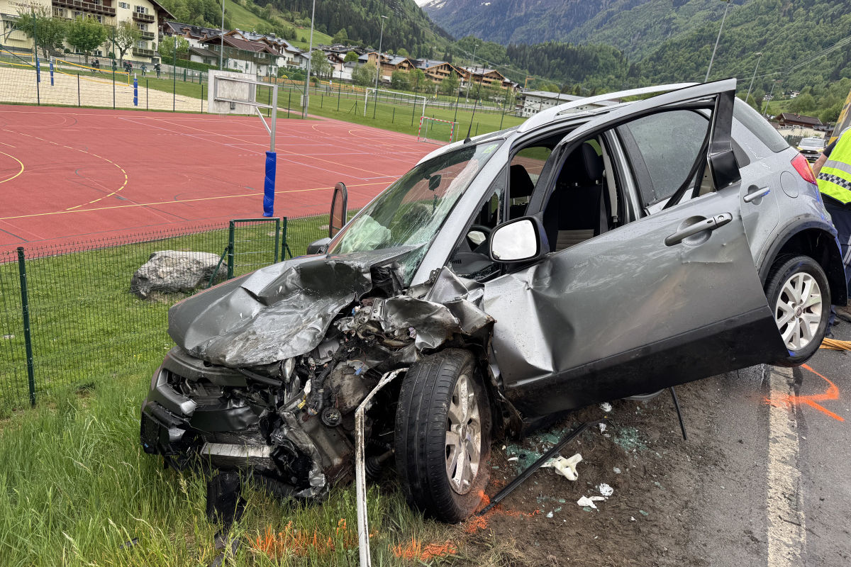 Ein stark beschädigtes Auto steht neben einem Sportplatz, mit erheblichem Schaden an der Front und der rechten Seite. Im Hintergrund sind grüne Hügel und ein Wohngebiet zu sehen.